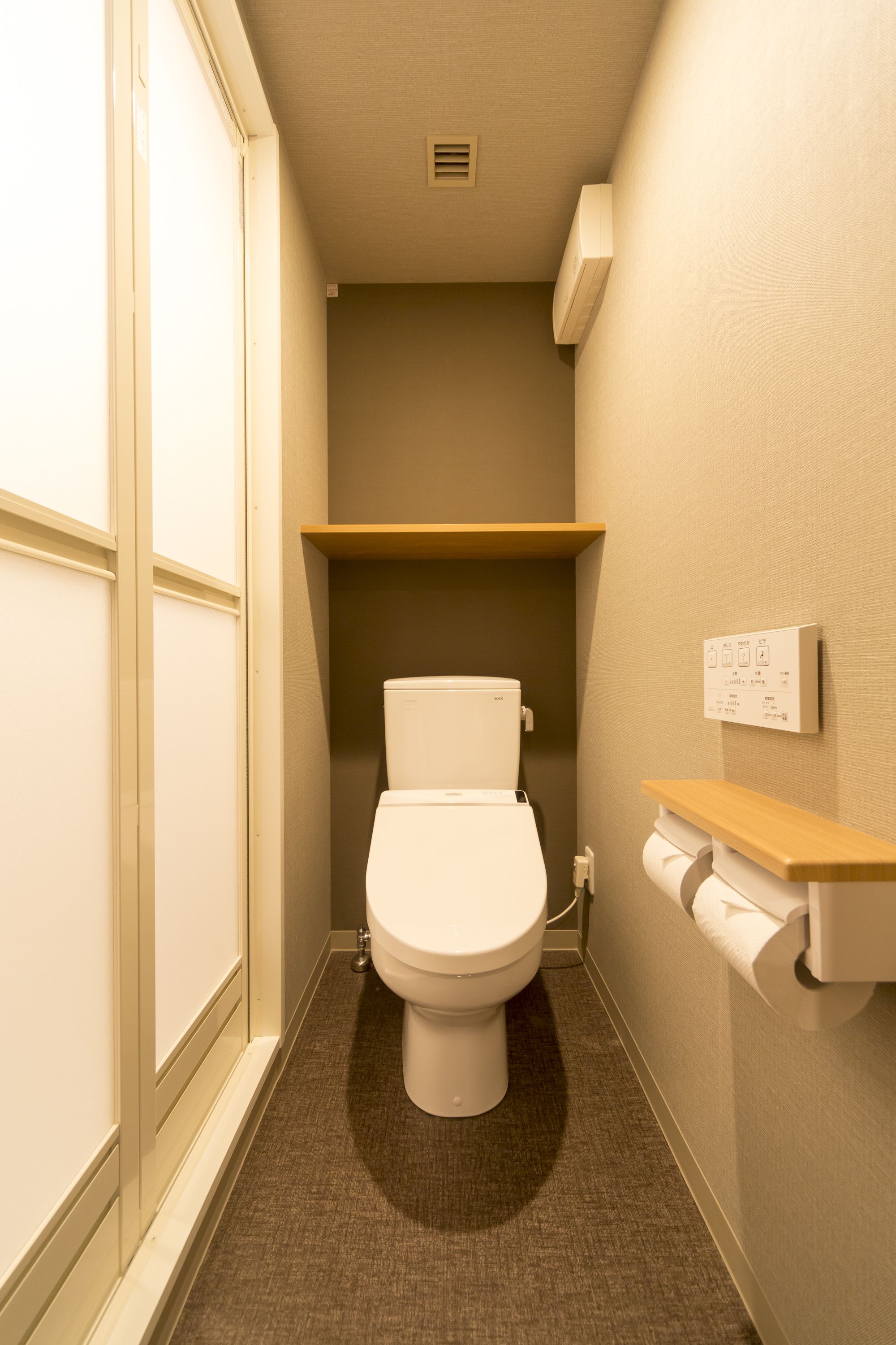 Une petite salle de bain comprenant des toilettes, une étagère en bois et un thermostat fixé au mur.