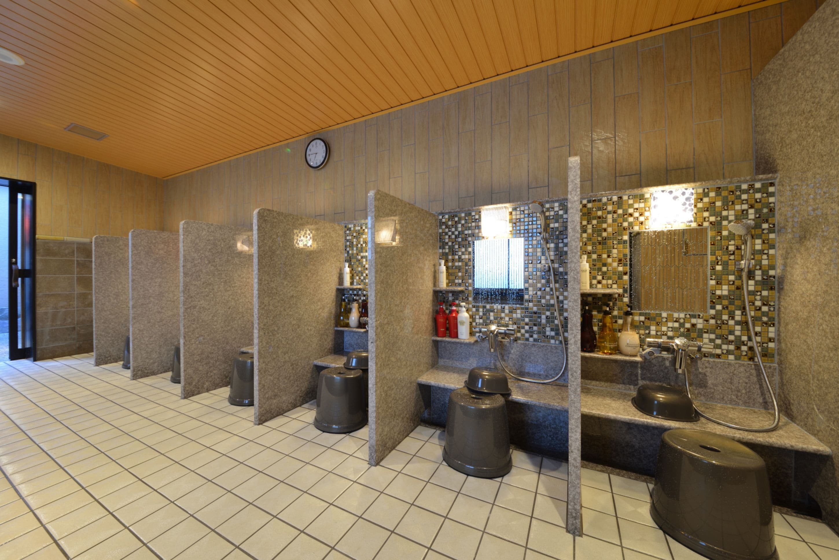 A public restroom with individual cubicles, a clock, and tiled walls.