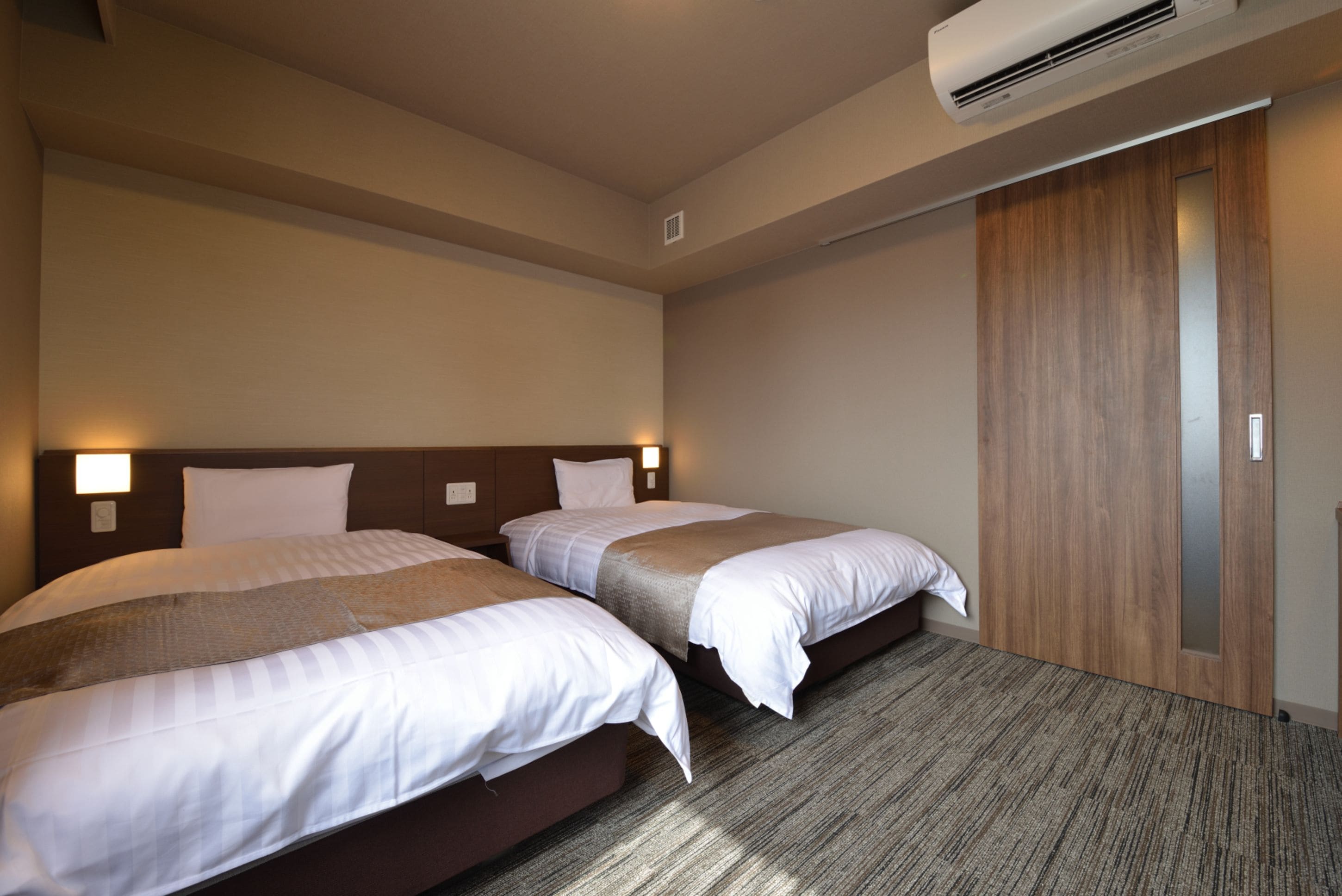 A hotel room with two beds, a wooden wardrobe, and an air conditioning unit.