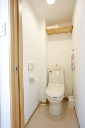 Casa de banho pequena e compacta com sanita, secador de mãos, dispensador de papel e prateleira sobre a sanita.