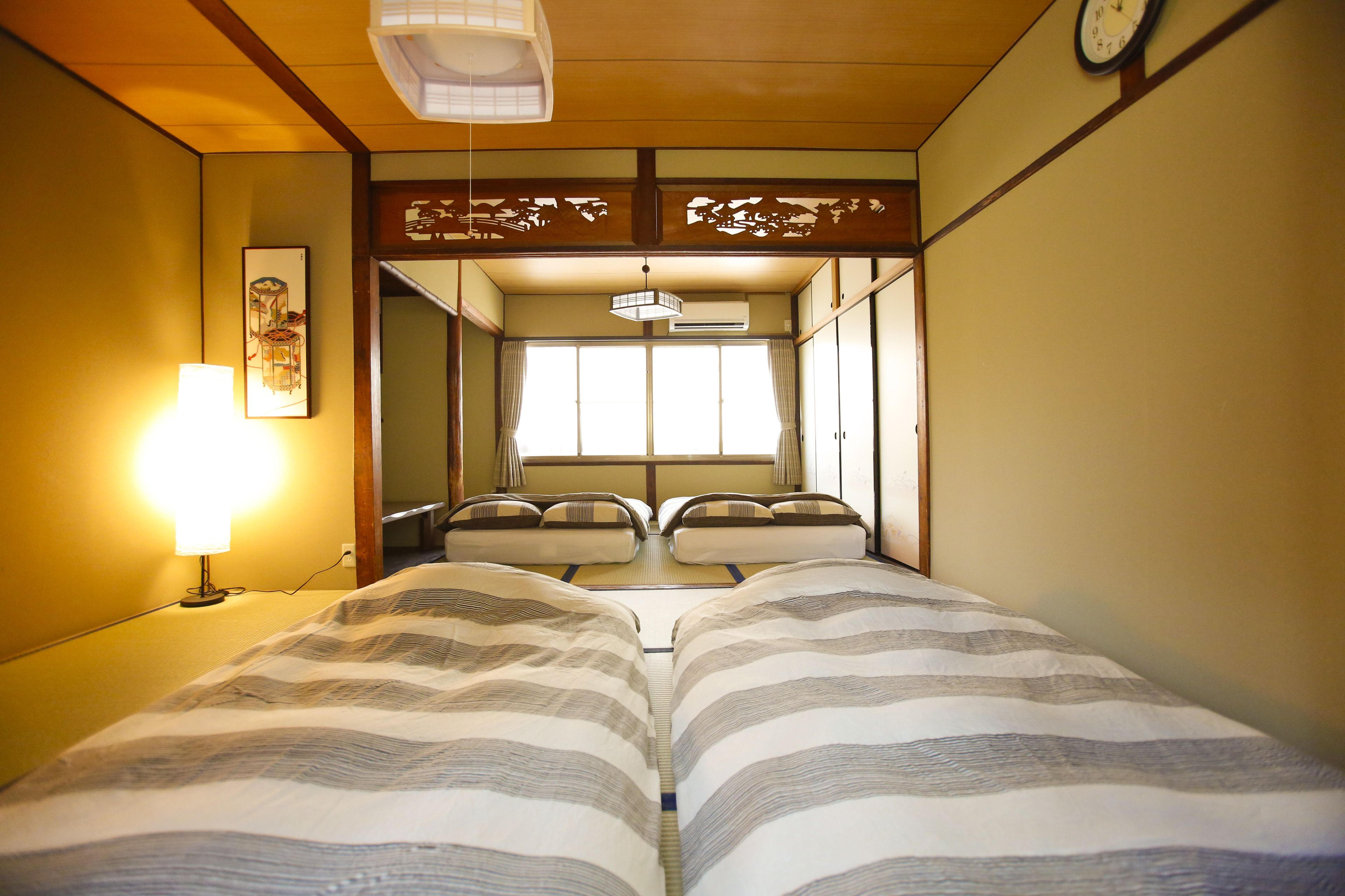 A traditional Japanese room with tatami flooring, a shoji screen, a bed with striped bedding, a bedside lamp, and a clock on the wall.