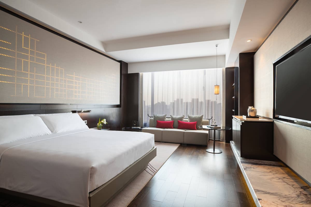 A modern hotel room with a large bed, a sofa, a small table, and a large window with a city view.