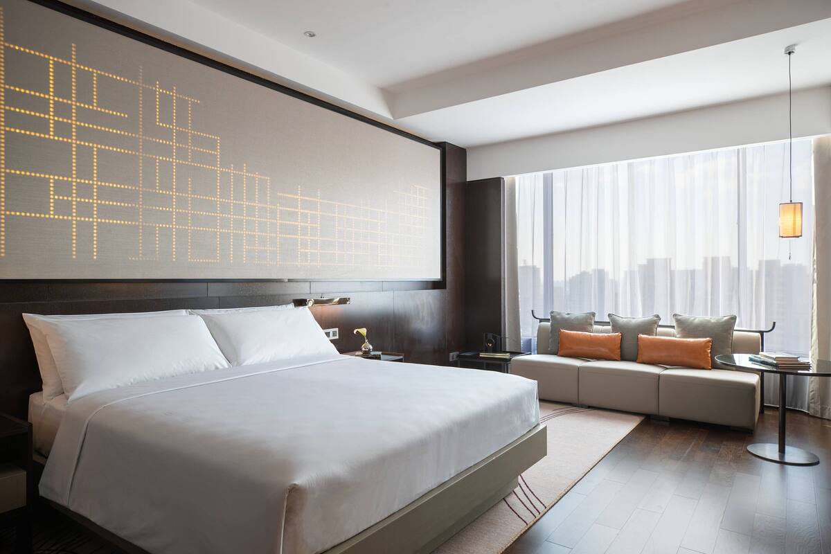 Una habitación de hotel moderna con una cama grande, un sofá, una mesita y vistas a la ciudad a través de amplios ventanales.