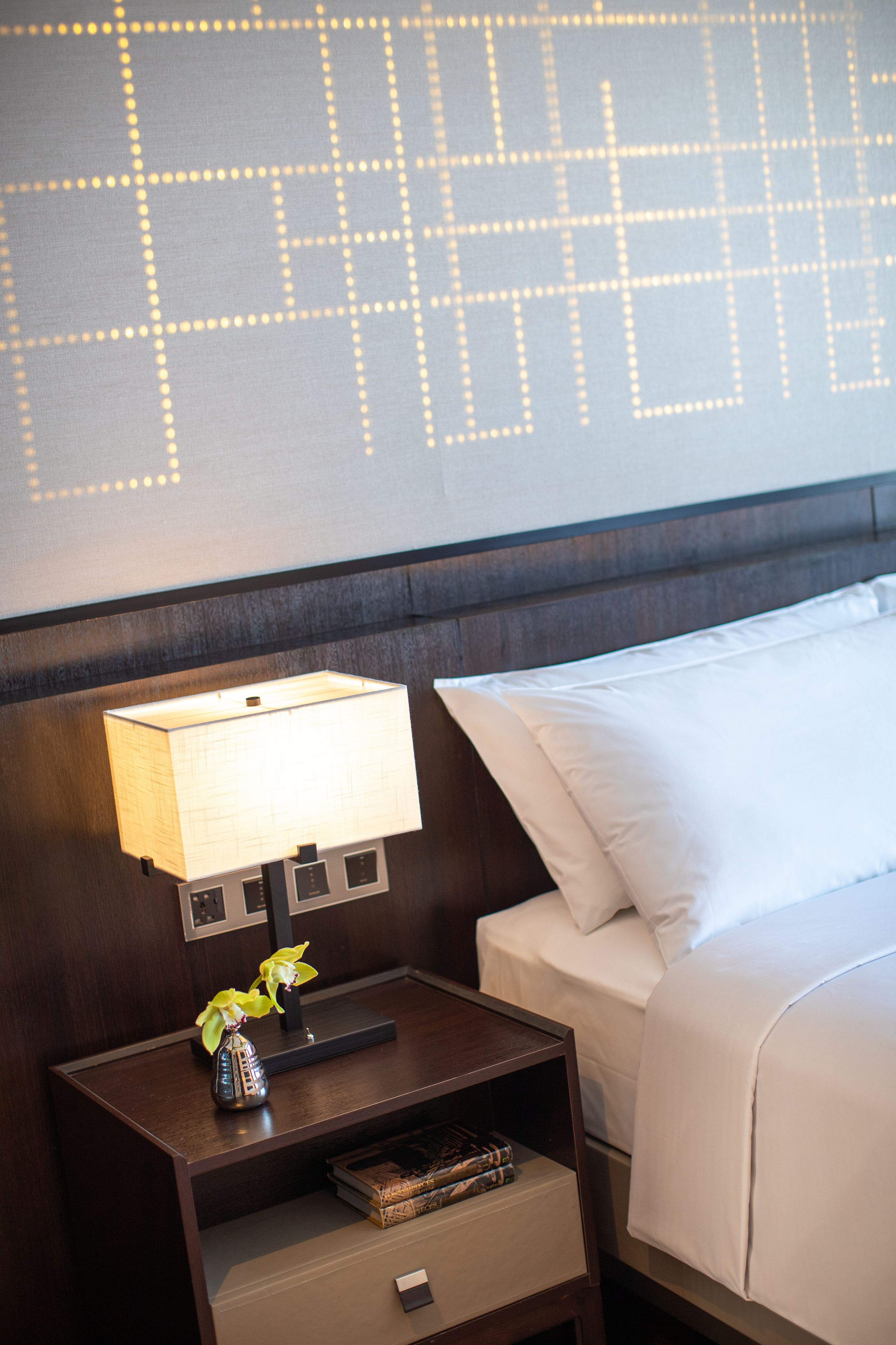 A neatly made bed with white linens, a bedside lamp, and a small nightstand with a vase of flowers and a book.