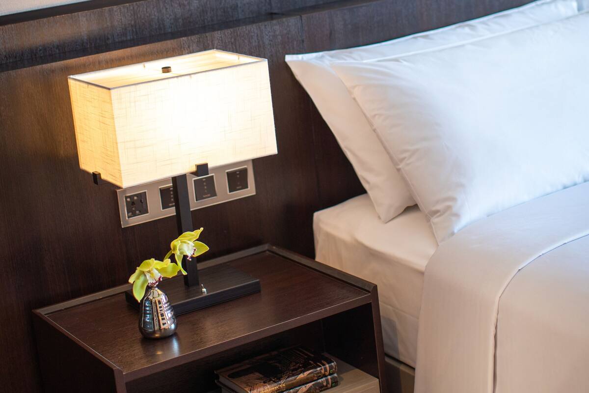 A neatly made bed with white linens, a bedside lamp, and a small nightstand with a vase of flowers and a book.