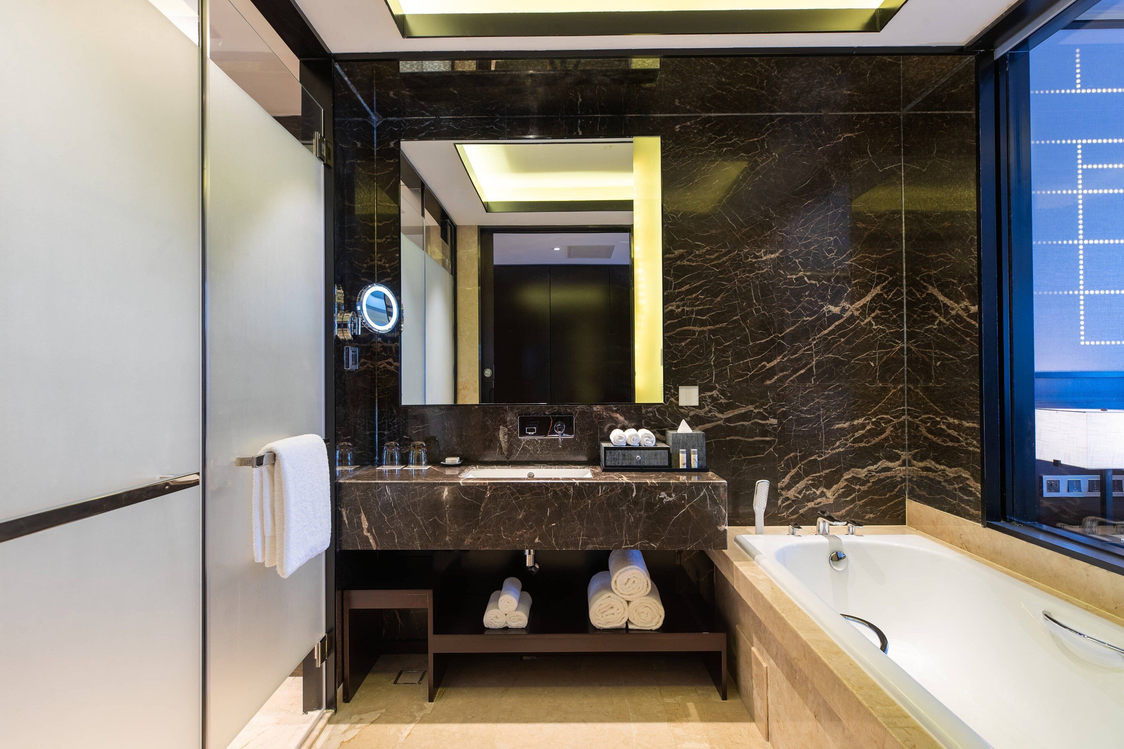 Un baño moderno con tocador de mármol, un espejo grande, una bañera y vistas al exterior.