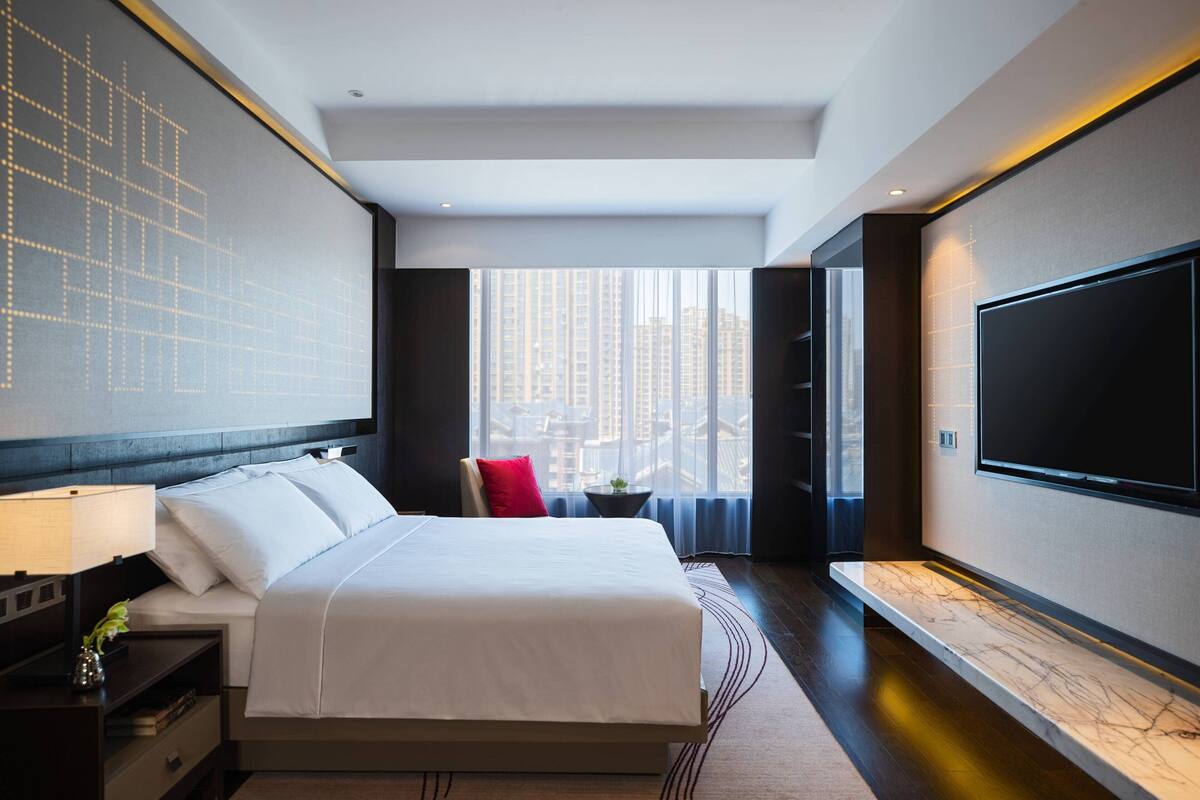 A modern hotel room with a large bed, a flat-screen TV, a desk with a lamp, and a view of tall buildings through the window.