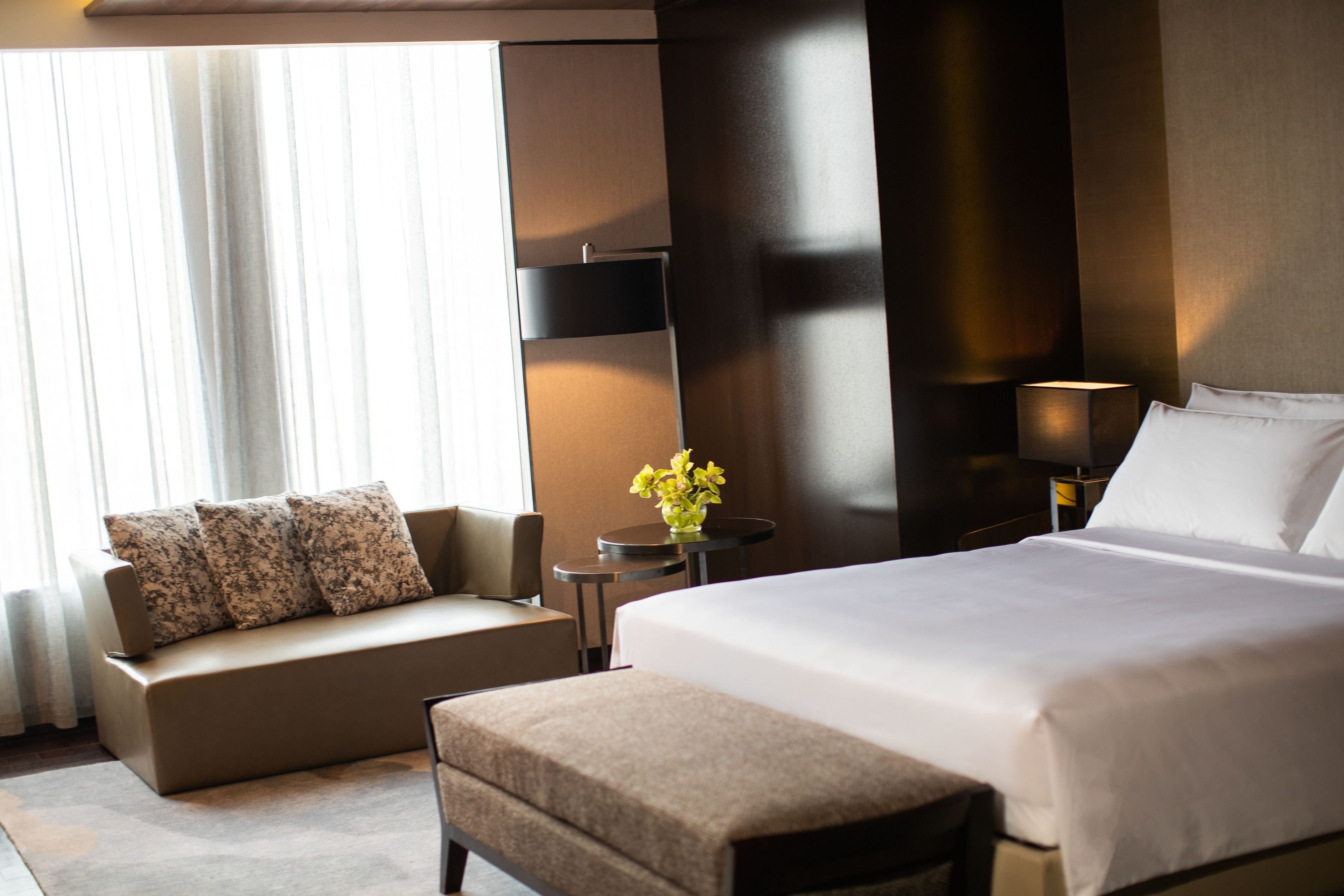 A modern hotel room with a large bed, a sofa, a small table with a vase of flowers, and a lamp.