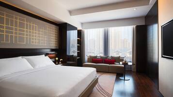 A modern hotel room with a large bed, a sofa, a small table, and a city view through large windows.