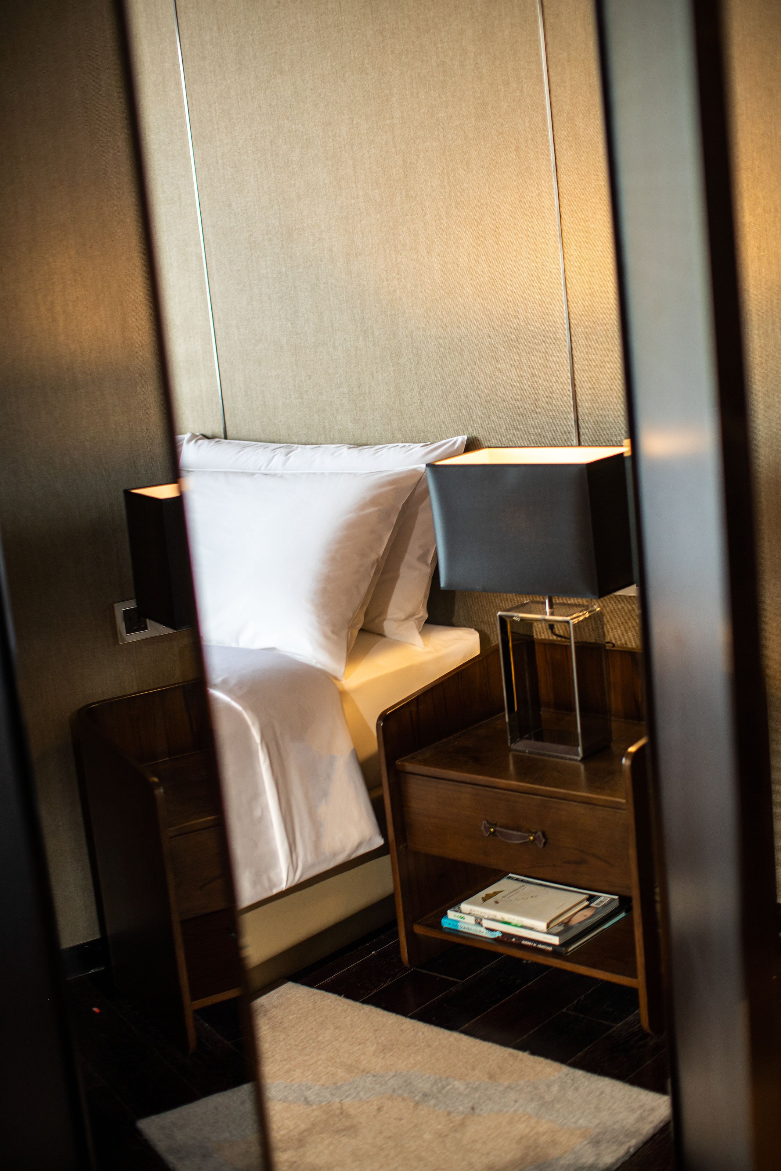 A hotel room with a bed, bedside table, lamp, and a small shelf with books.