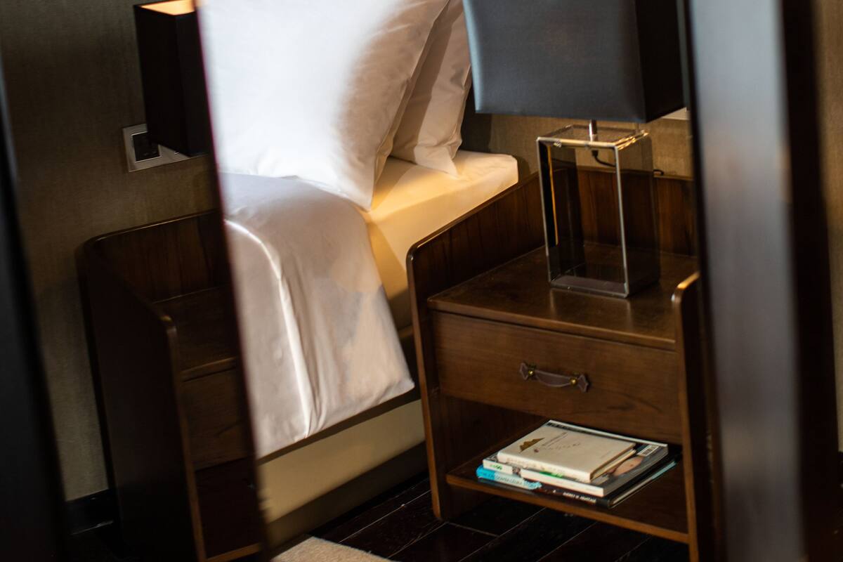 A hotel room with a bed, bedside table, lamp, and a small shelf with books.