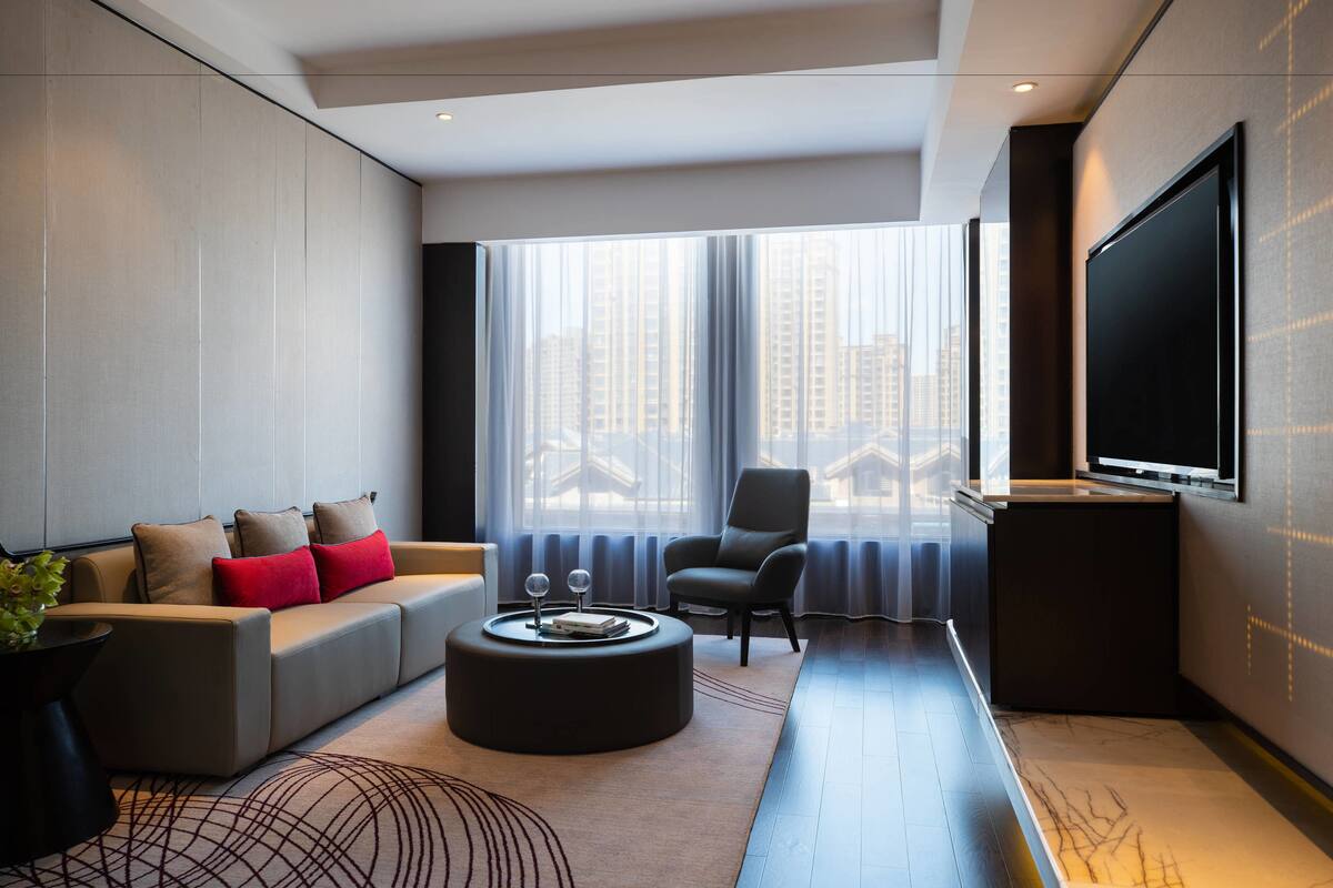 A modern living room with a sofa, ottoman, and chair, a large TV, and a window with a city view.