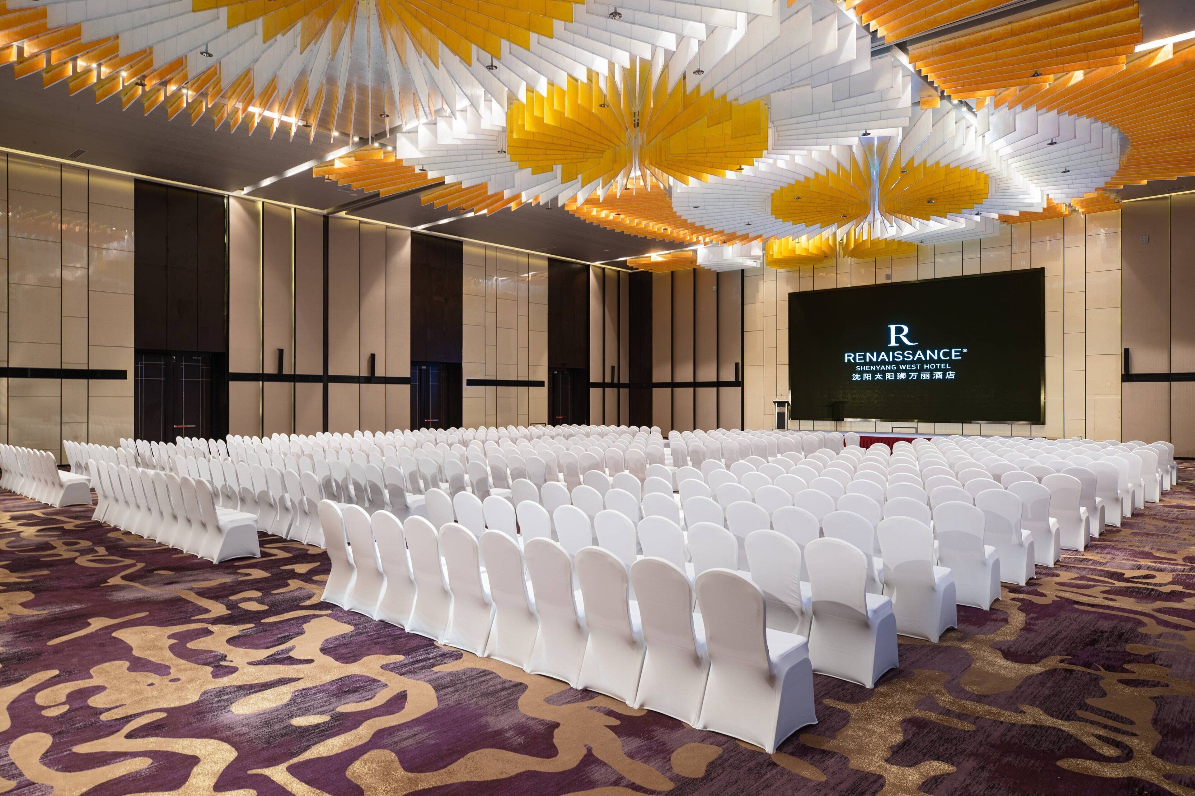 Un salón de conferencias con filas de sillas blancas, una pantalla grande que muestra el nombre del hotel y una alfombra estampada.