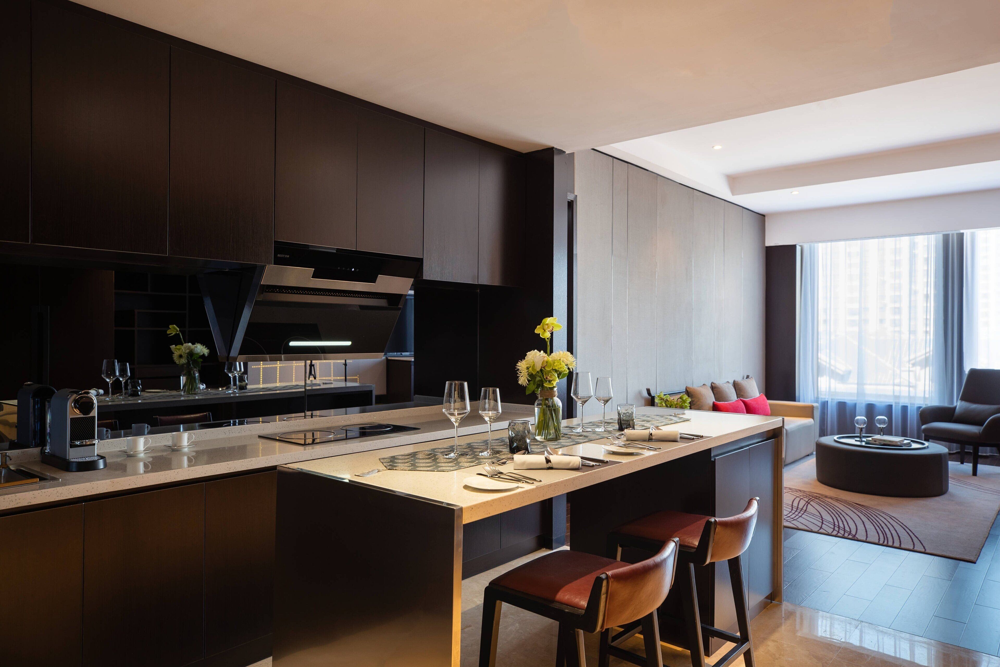 A modern kitchen with dark cabinetry, a central island with bar stools, and a dining area with a sofa and coffee table.