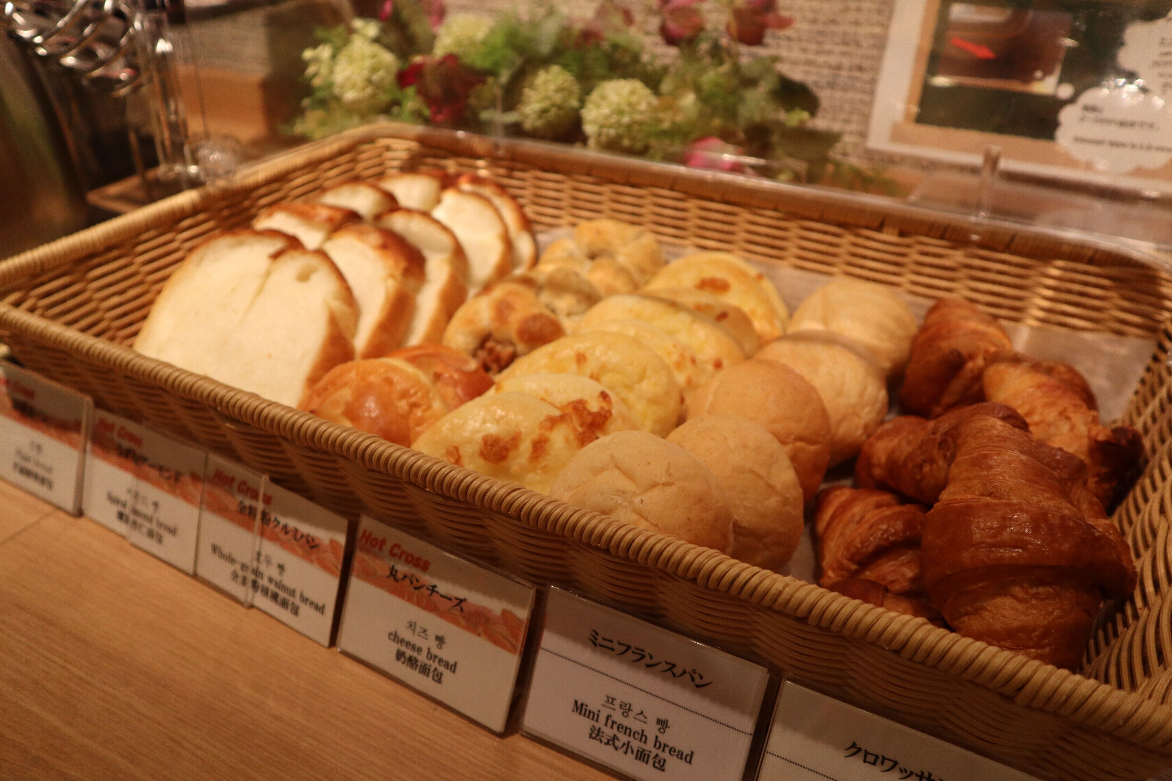 A basket of various breads and pastries, including whole grain and mini french bread, displayed in a bakery setting.
