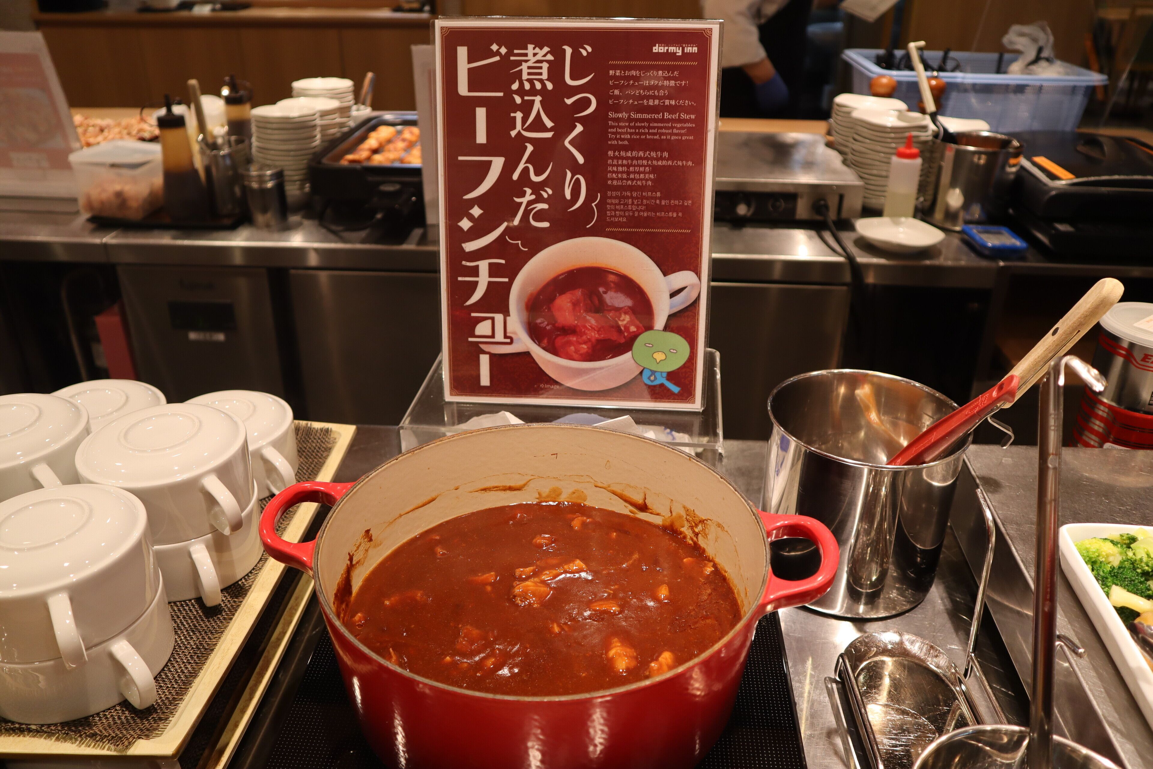 Un comptoir de cuisine avec une marmite rouge contenant un ragoût, des bols blancs et une pancarte avec du texte en japonais.