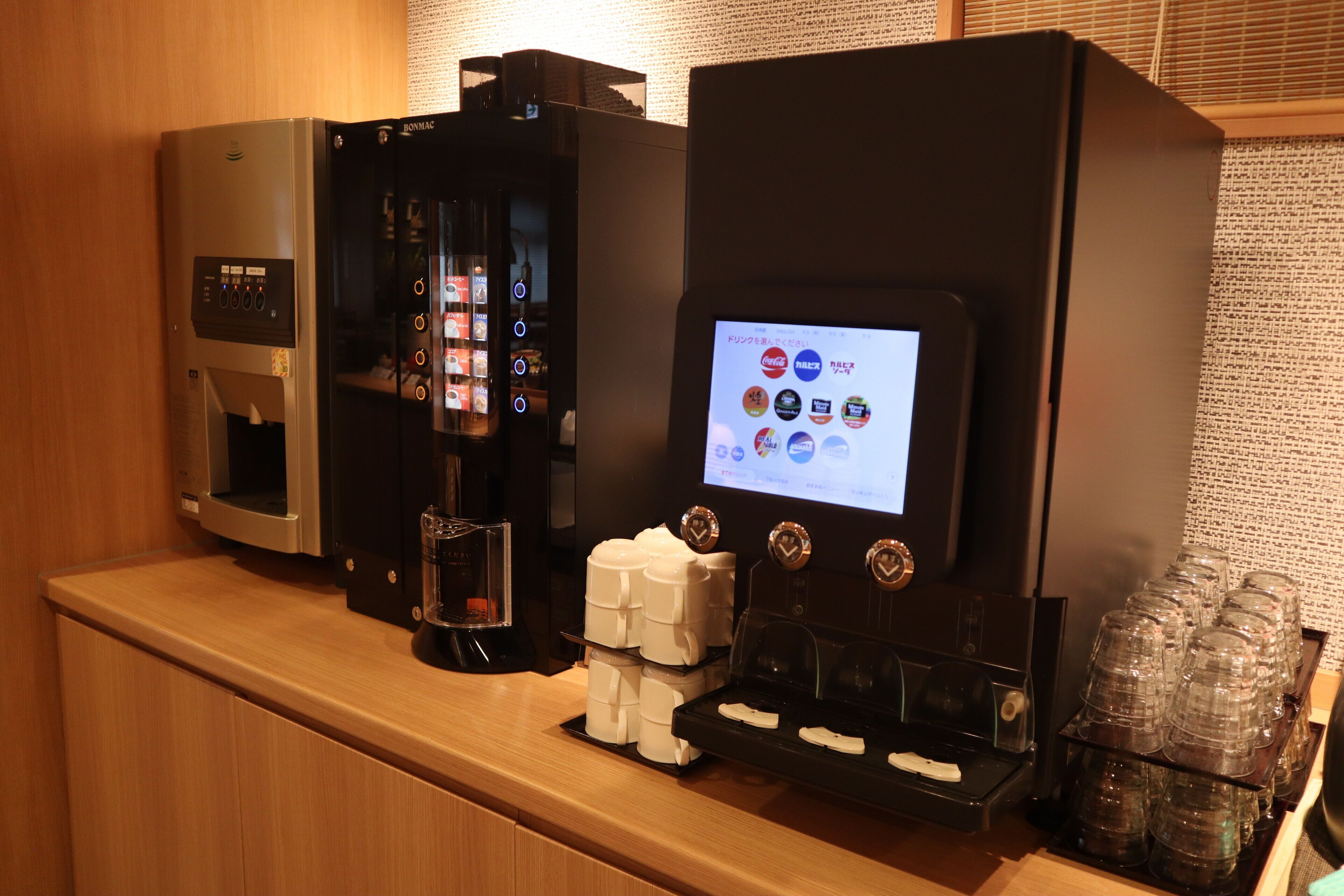 Una macchina da caffè con display digitale, caraffa in vetro e una pila di bicchieri di carta su un bancone di legno.