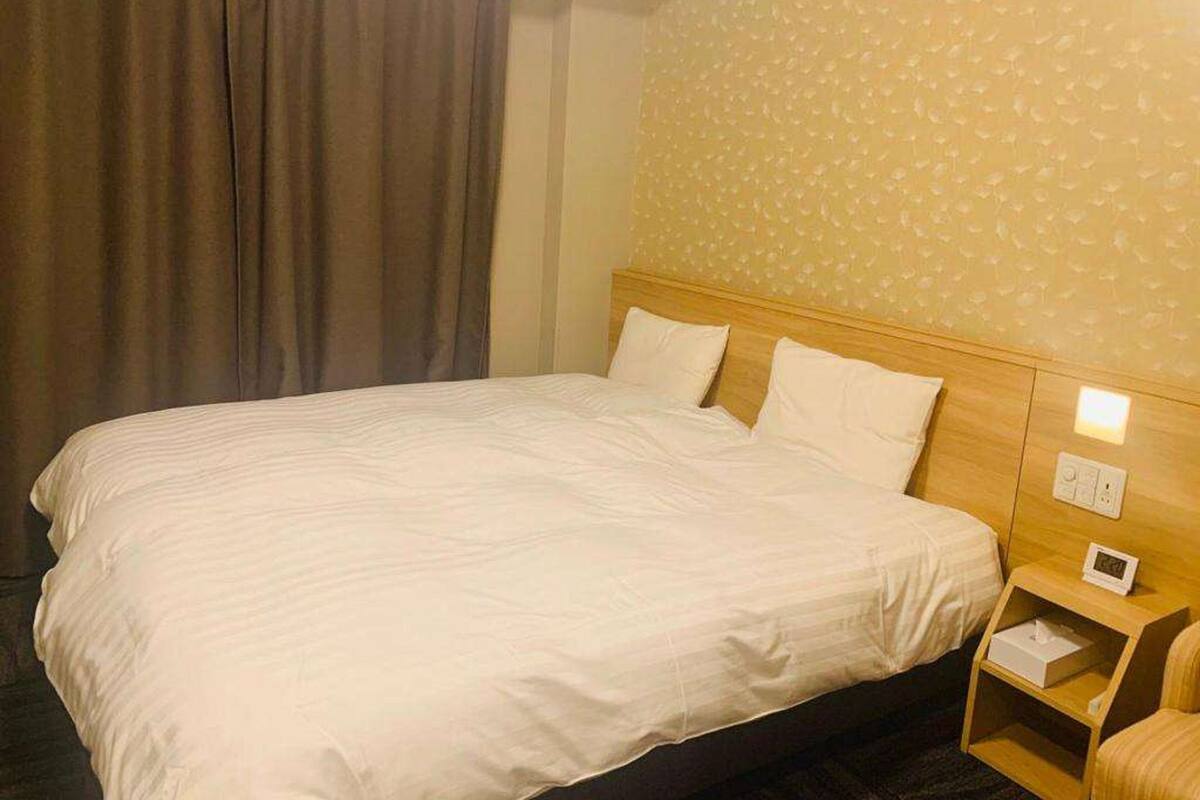 A hotel room with a large bed, a wooden headboard, a nightstand with a lamp, and a window with curtains.