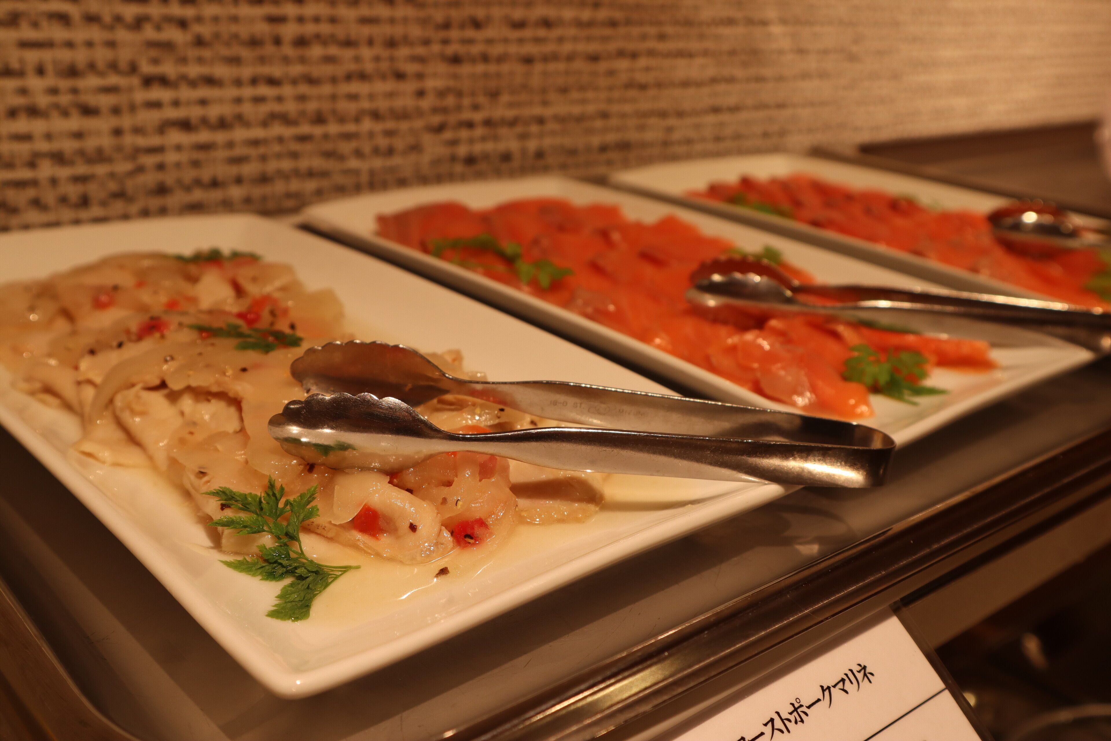 A buffet with various dishes, including seafood and a creamy pasta dish, served on white plates with tongs.