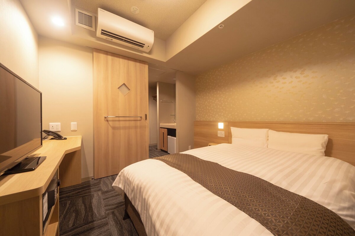 A hotel room with a bed, a desk with a phone, an air conditioner, and a wall-mounted light.