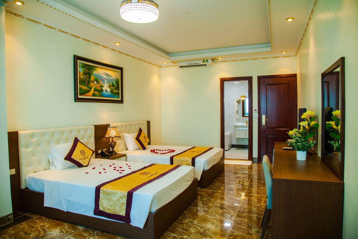 A hotel room with two beds, a painting on the wall, an air conditioning unit, and a desk with a vase of flowers.