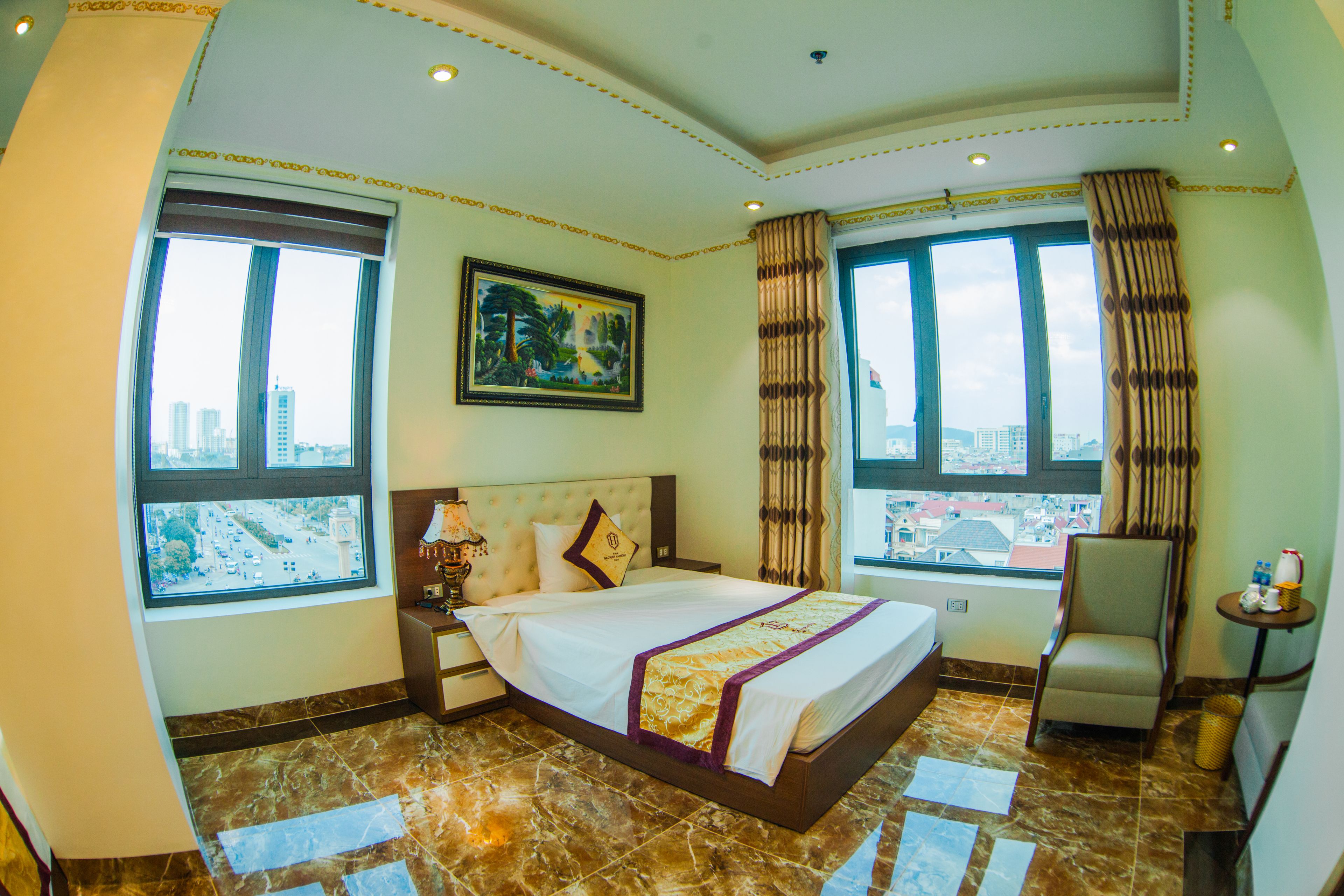 A hotel room with a large bed, two windows, a painting, a chair, and a small table.