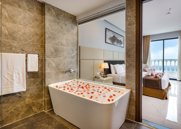 A modern bathroom with a large bathtub, marble walls, a visible bed with a view, and a framed picture on the wall. - Imperial Nha Trang Hotel (Nha Trang)