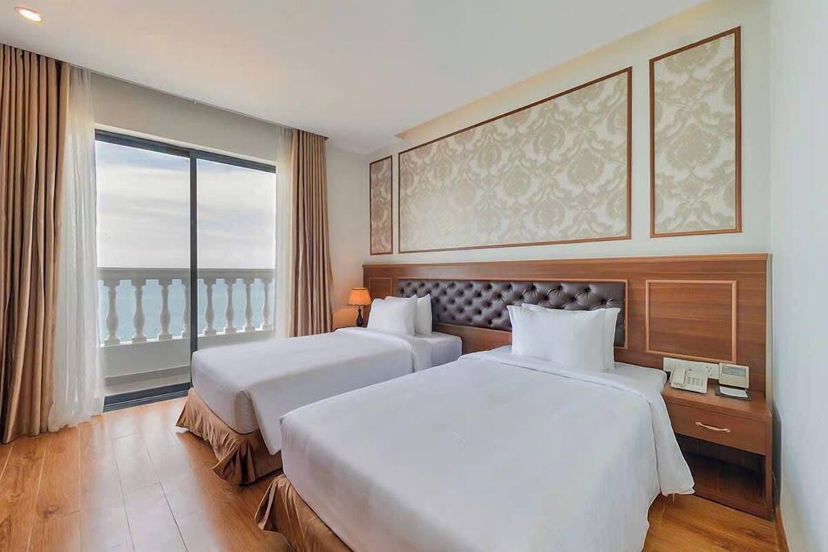 A hotel room with two beds, a large window with a view, a bedside table with a lamp, and a decorative wall panel.