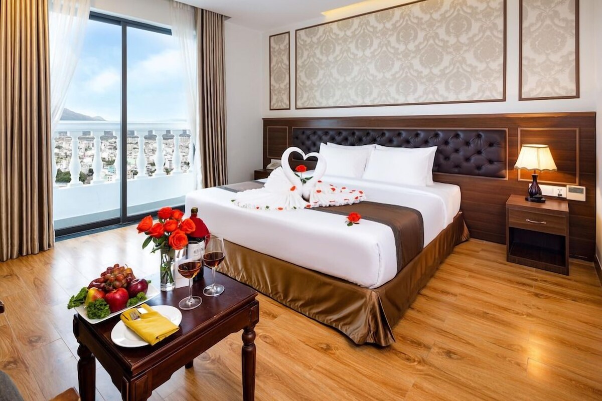 A hotel room with a large bed, a view of the city, a dining table with fruits and wine, and a nightstand with a lamp.