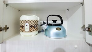 A kitchen cabinet with a white interior, containing a white electric kettle with a black handle and a patterned rice cooker. - Studio at Green Pramuka City Serene Apartment Living By Travelio (Jakarta)