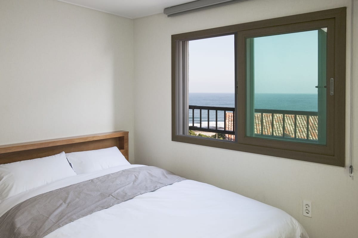 A bedroom with a bed, a window with a view of the sea, and a balcony with a railing.
