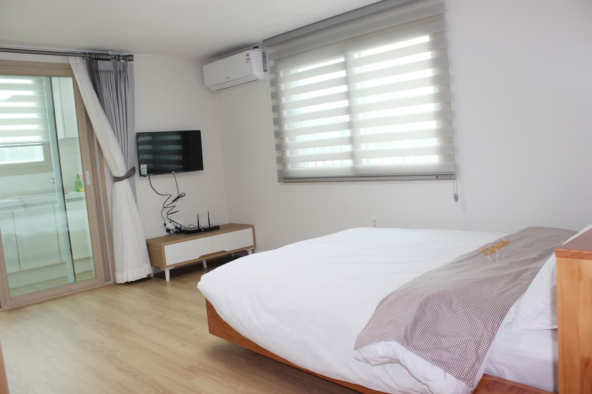 A bedroom with a bed, a TV, a window with blinds, a bedside table, and a sliding glass door.
