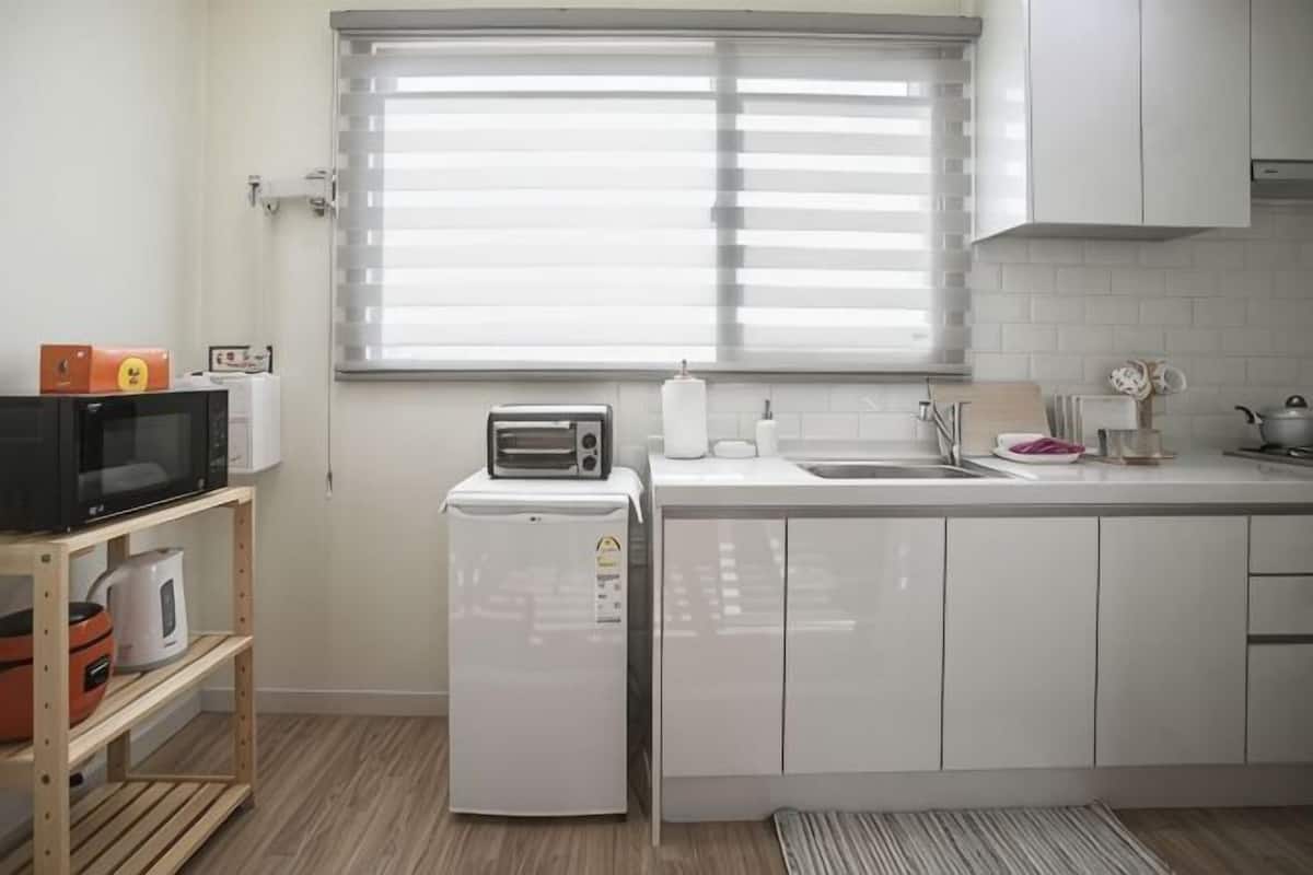 A kitchen with a microwave, toaster, and refrigerator, a window with blinds, and white cabinets.