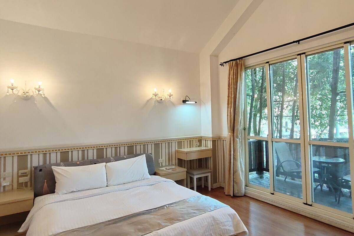 A spacious bedroom with a large bed, a chandelier, a desk, and a view of the outdoors through large windows.
