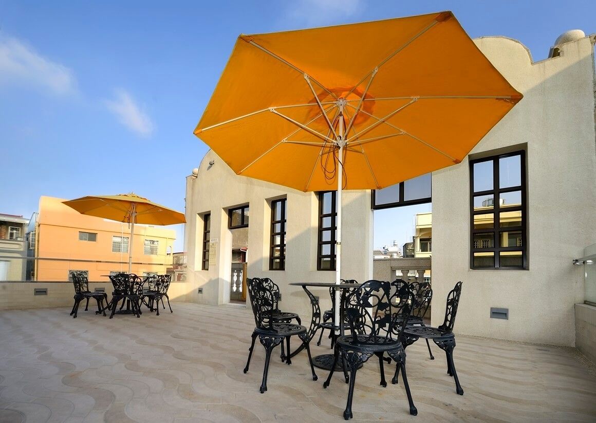Un patio al aire libre con mesas y sillas, sombrillas grandes y un edificio con ventanas y puertas.