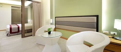 Una habitación de hotel con una cama, una mesita redonda de vidrio con una planta, una silla de mimbre blanca y una mesita de noche con una lámpara.