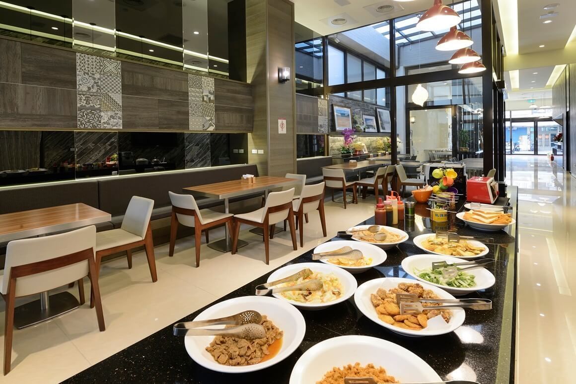 A modern restaurant with a long counter displaying various dishes, surrounded by tables and chairs.