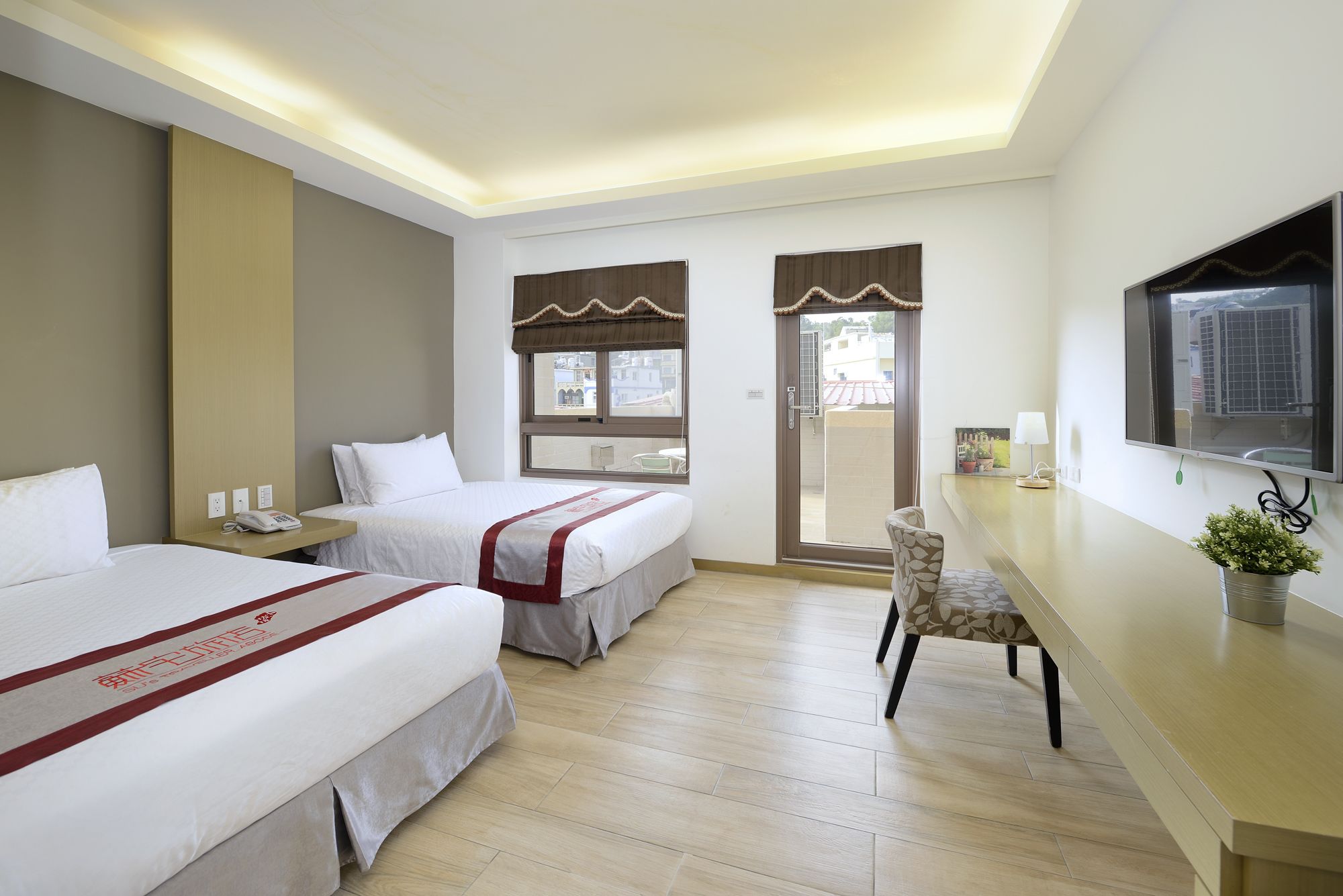 A hotel room with two beds, a desk, a chair, a TV, and a balcony with a view.