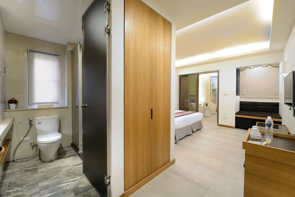 A modern hotel room with a wooden wardrobe, a bed with a red blanket, a small desk with a water bottle, and a bathroom with a toilet and shower.