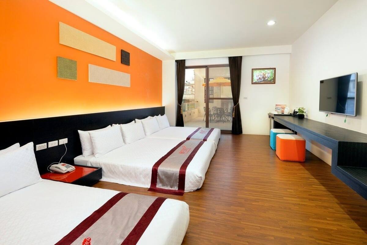 A hotel room with two beds, a TV, a desk, and a balcony with outdoor seating.