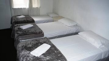 Une chambre d’hôtel avec trois lits simples, chacun agrémenté d’un oreiller blanc et d’une serviette blanche pliée. Les lits sont recouverts de draps gris et blancs.
