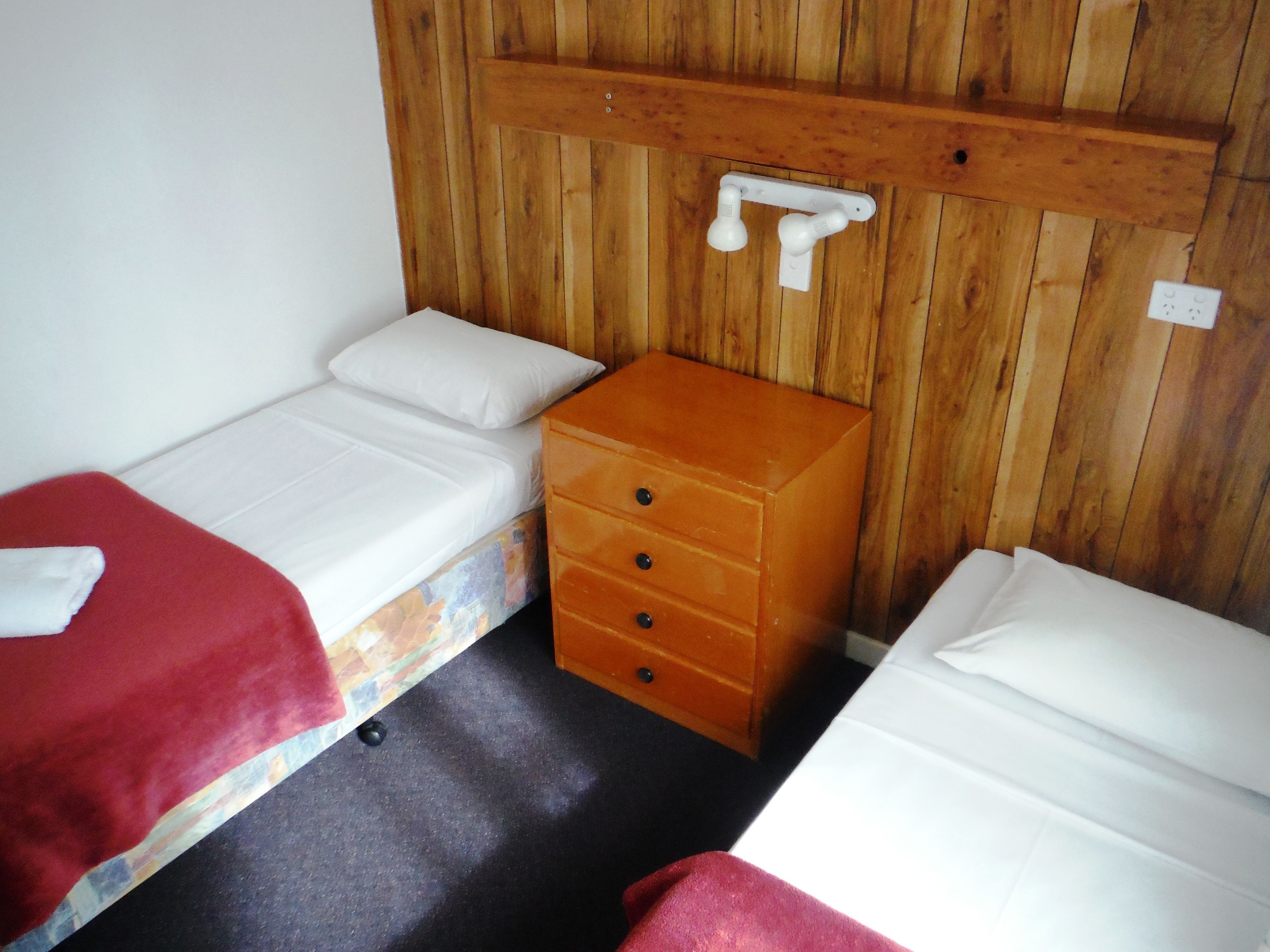 A small, compact room with two single beds, a wooden dresser, and a wooden wall panel.