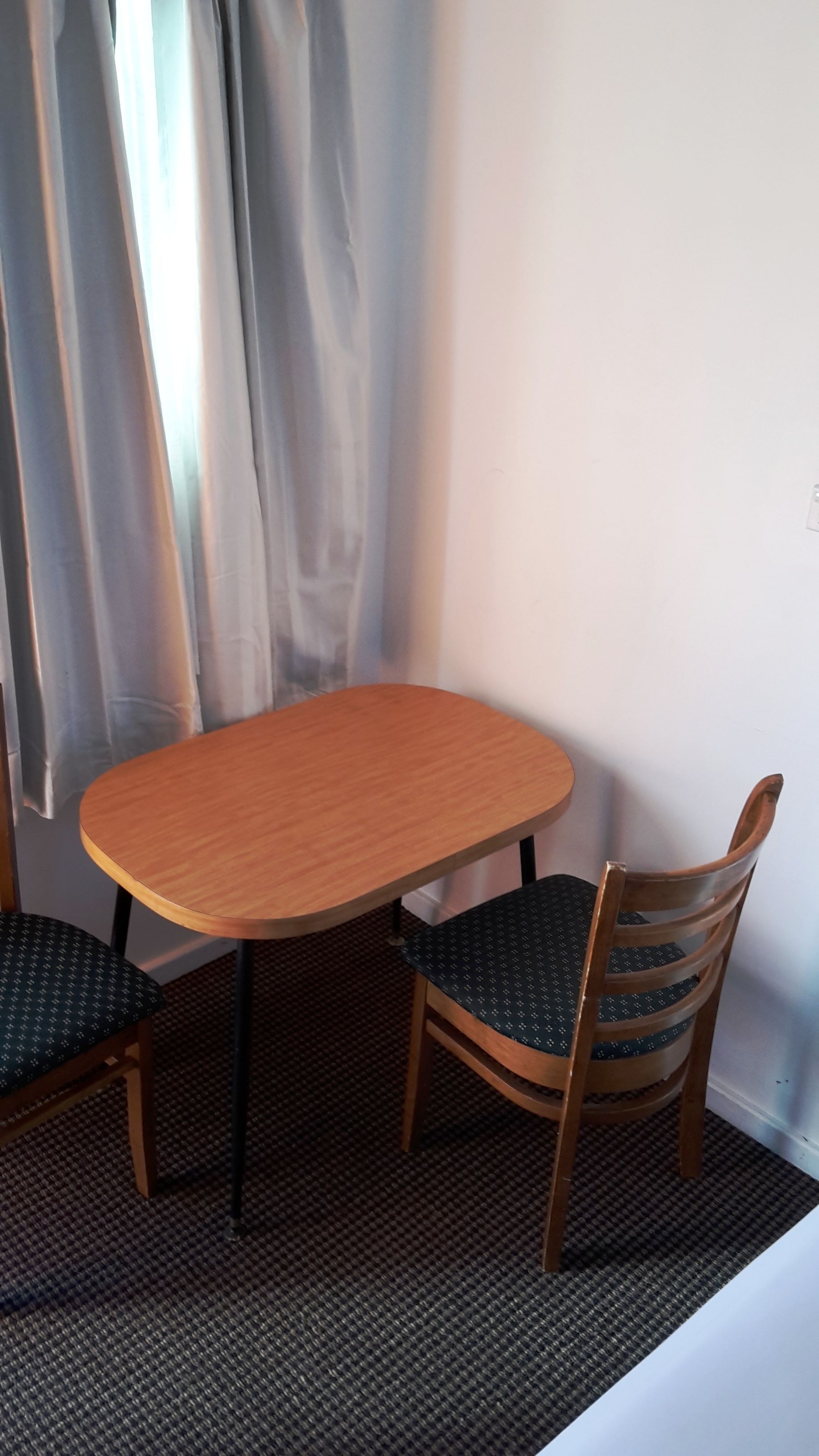 A small dining area with a wooden table and two chairs, a patterned carpet, and a window with curtains.