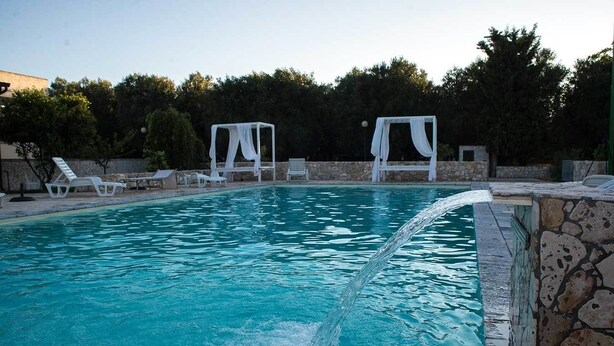 Una piscina con agua azul cristalina, sillas de descanso blancas y un muro de piedra.