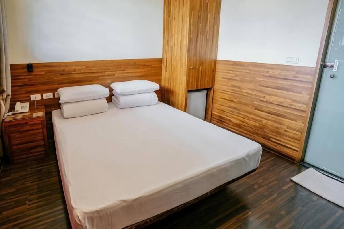 Habitación de hotel con cabecera de madera, cama con ropa blanca, mesita de noche con teléfono y puerta al baño.