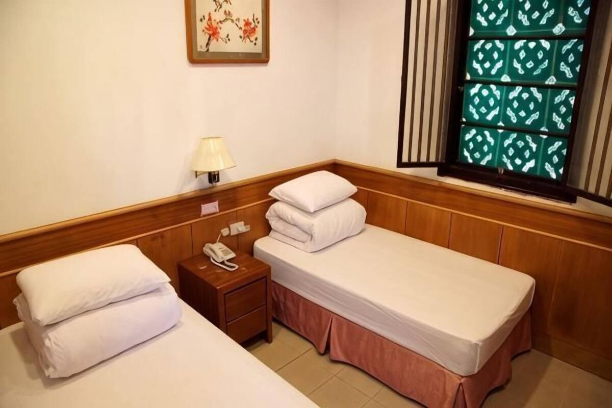 Habitación de hotel con dos camas, una mesita de noche con lámpara, teléfono y ventana con cortinas estampadas.