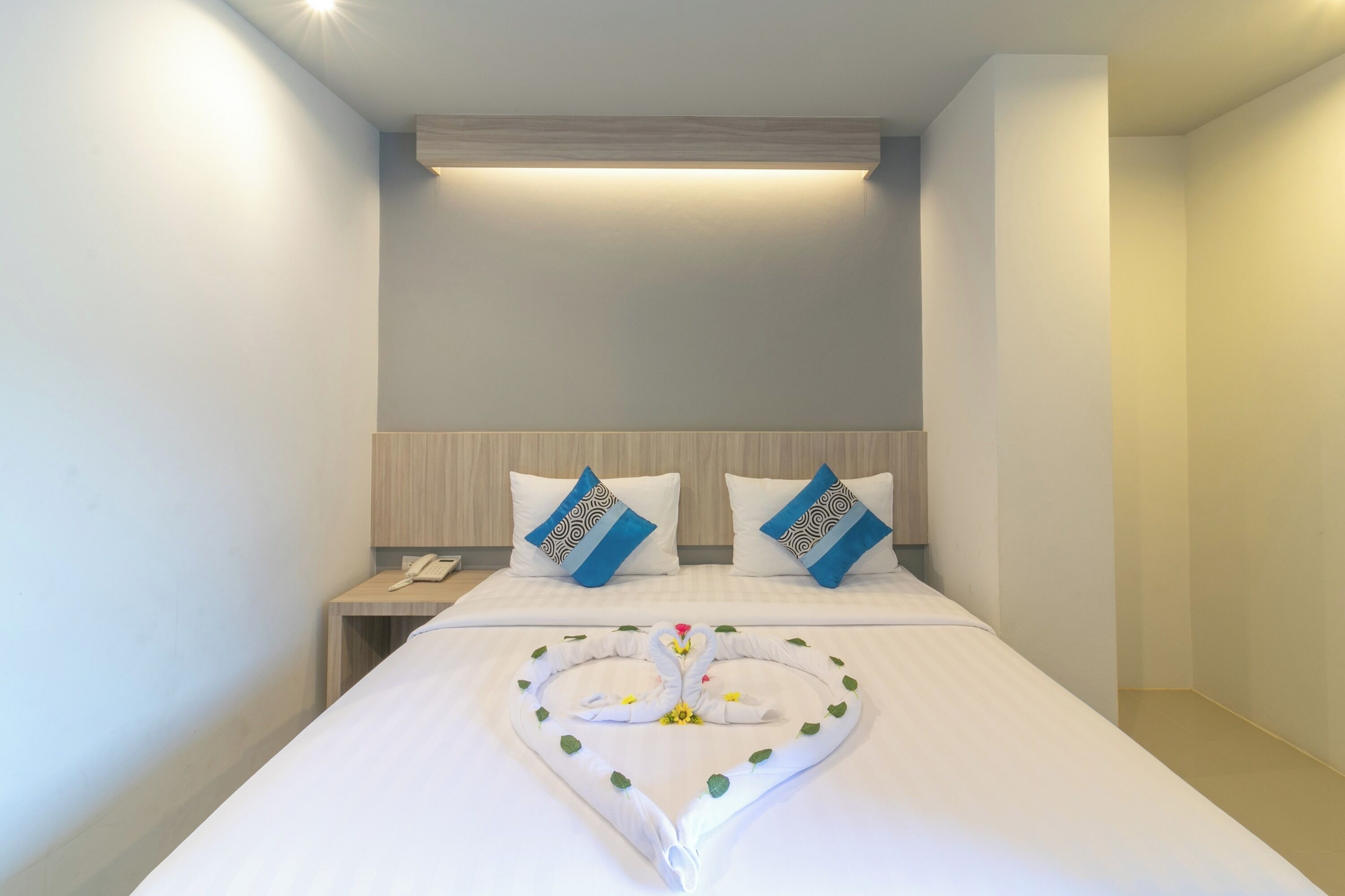A hotel room with a neatly made bed, two pillows with a blue and white pattern, a bedside table with a phone, and a heart-shaped decoration on the bed.