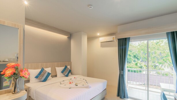 A hotel room with a large bed, a bedside table with a vase of flowers, a window with curtains, and a balcony. - Carpio Hotel Phuket (Wichit)