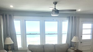 Una sala de estar con sofá, sillón y mesa de centro, ventanas amplias con vista al mar y ventiladores de techo.