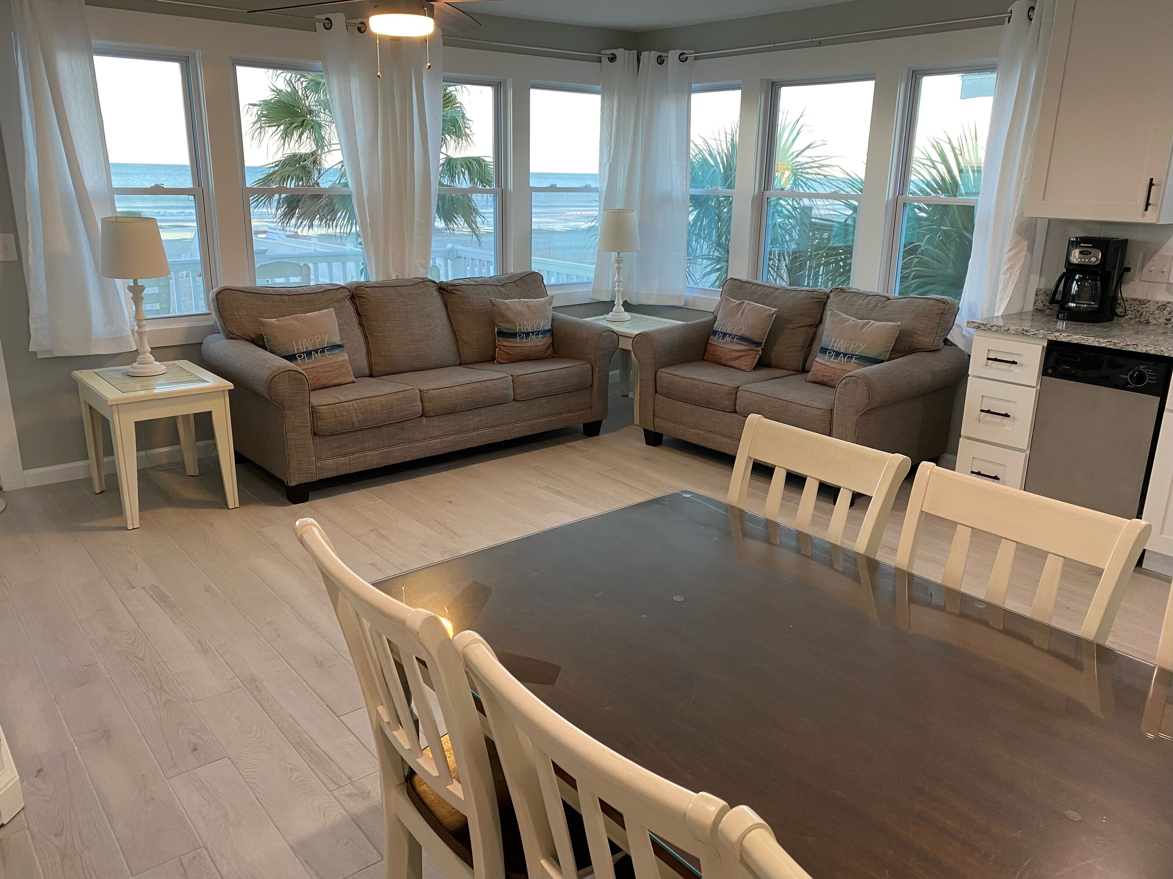 Ein Wohnzimmer mit zwei Sofas, einem Esstisch und Stühlen. Große Fenster mit Vorhängen, eine Lampe auf einem Beistelltisch und eine Kaffeemaschine auf der Arbeitsfläche.